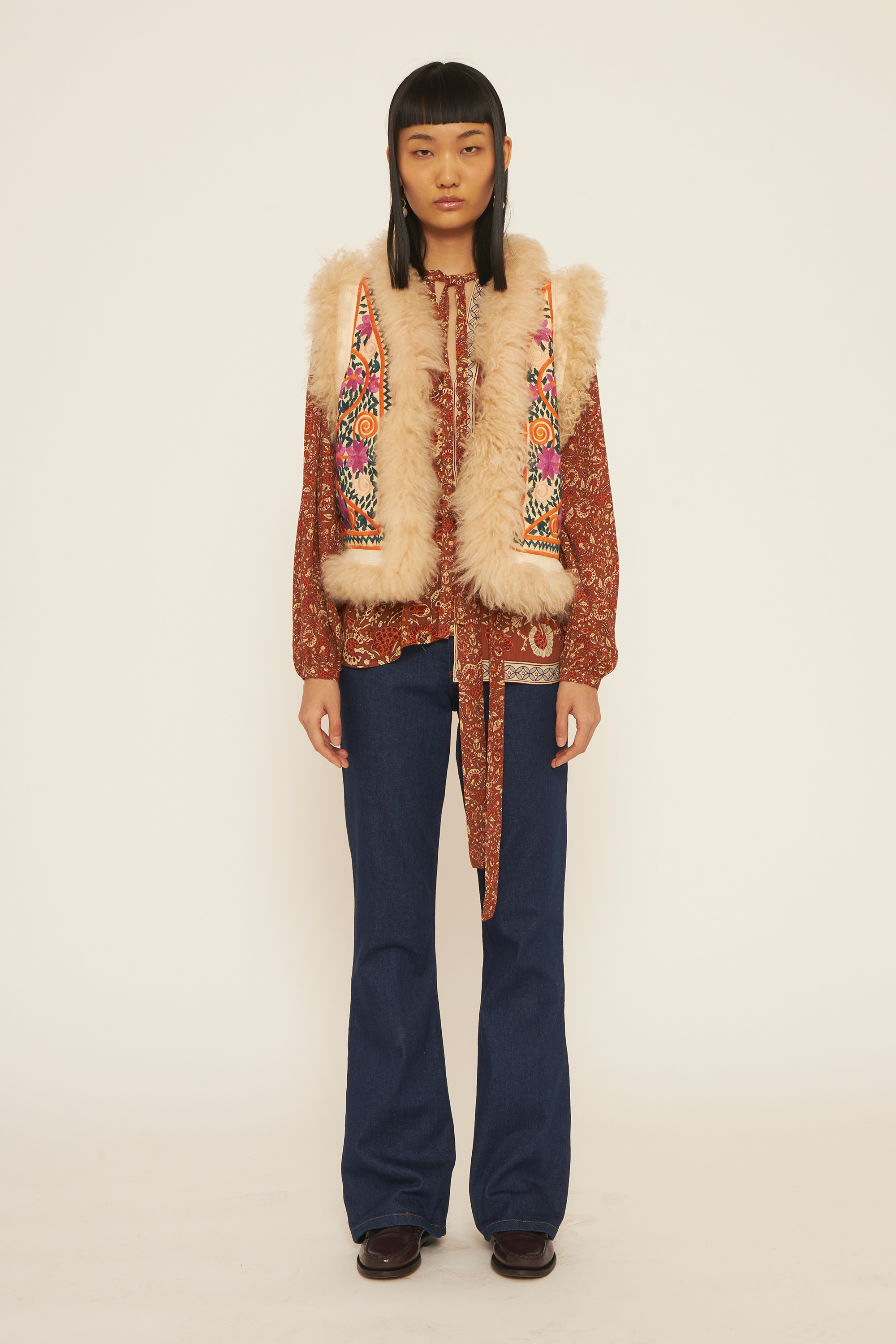 A girl wearing a rust blouse, a pair of jeans and a colorful embroidery gilet.