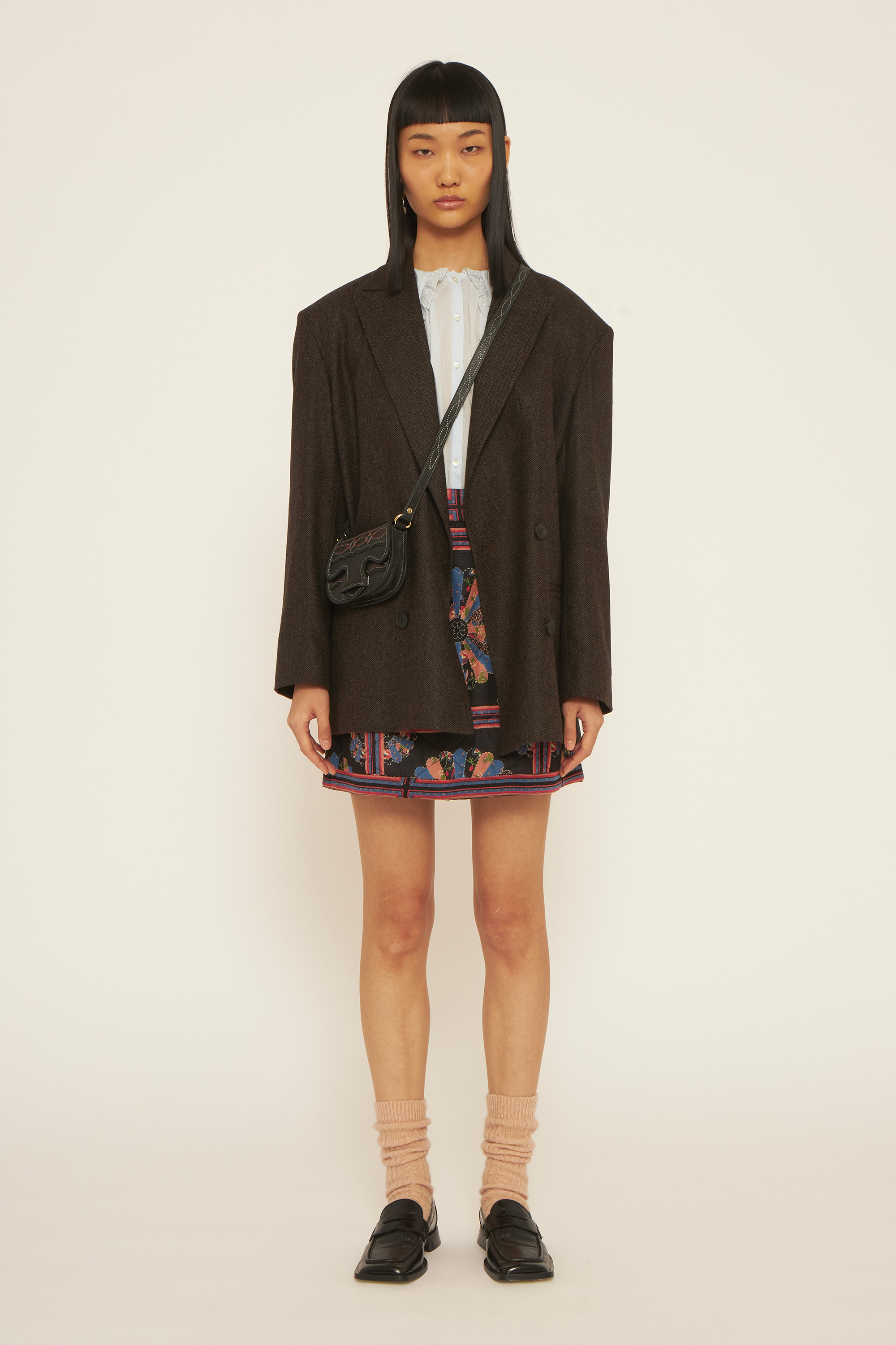 A girl wearing a brown jacket, a light blue delicate blouse, a printed skirt and a black bag.